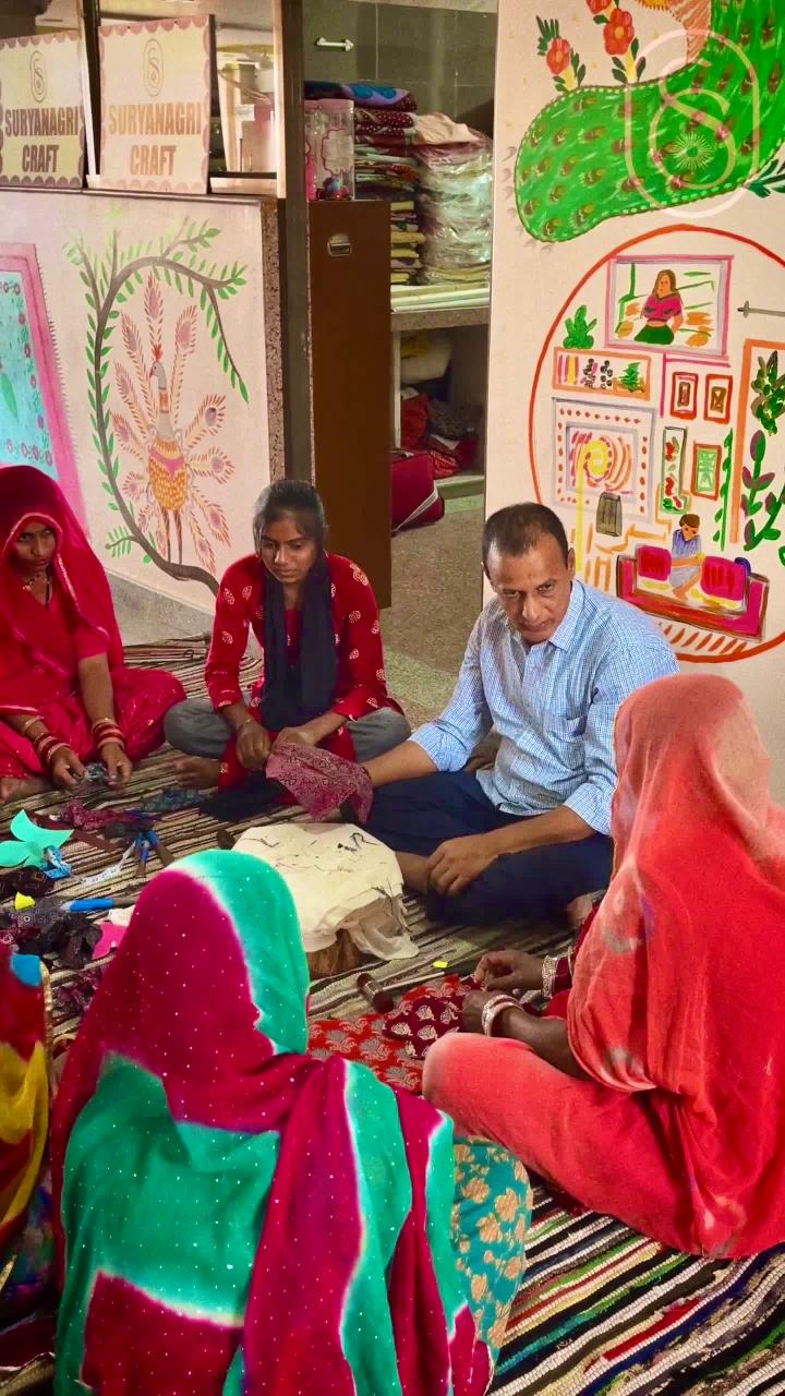 Group of people sitting on the floor in a room with colorful murals and art supplies discussing embroidery
